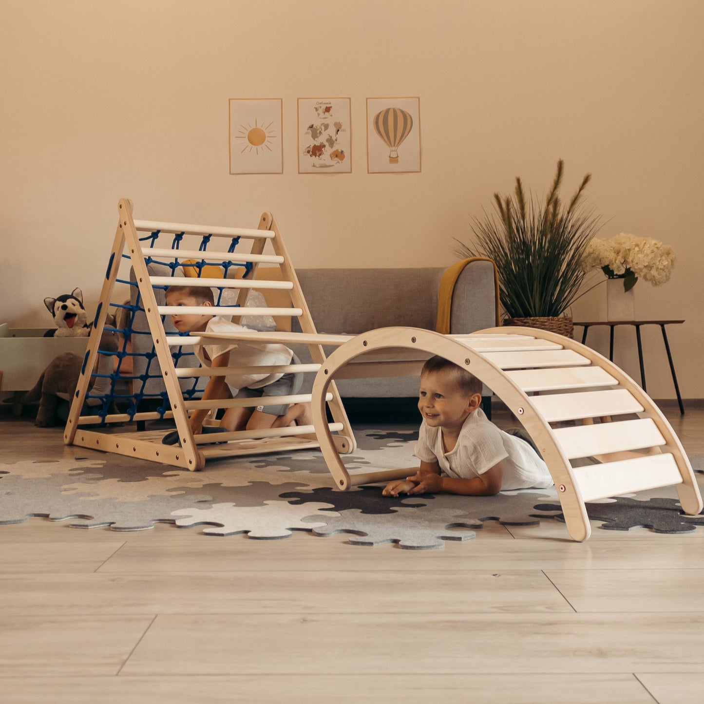 Zwei Kinder spielen im Kinderzimmer mit einem Kletterset aus Holz, bestehend aus einem dreiseitigen Kletterdreieck mit Netz, einer Rutschrampe und einem Kletterbogen, der als Wippe genutzt wird.