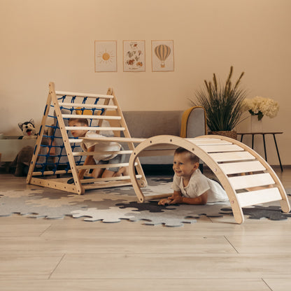 Zwei Kinder spielen im Kinderzimmer mit einem Kletterset aus Holz, bestehend aus einem dreiseitigen Kletterdreieck mit Netz, einer Rutschrampe und einem Kletterbogen, der als Wippe genutzt wird.